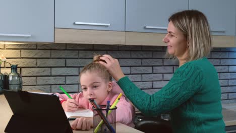 Madre-caucásica-está-arreglando-el-pelo-de-su-hija-mientras-ella-está-teniendo-clases-en-línea-usando-una-computadora-portátil