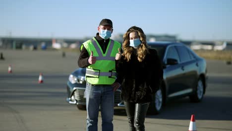 Mujer-y-hombre-caucásicos-confiados-con-máscaras-faciales-covid-que-muestran-pulgares-hacia-arriba-con-el-coche-en-el-fondo.-Retrato-del-instructor-de-conducción-y-estudiante-posando-sobre-la-pandemia-de-coronavirus-al-aire-libre