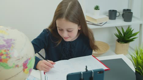 Distance-learning-concept.-Schoolgirl-studying-online-using-smartphone-making-notes-in-copybook.-Teen-girl-school-student-watching-internet-video-course-sitting-at-home-desk.