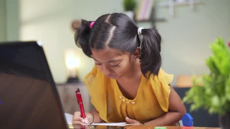 Concept-of-homeschooling,-online-education-or-e-learning,-young-girl-busy-in-writing-by-looking-into-laptop-while-teacher-explaining-during-class