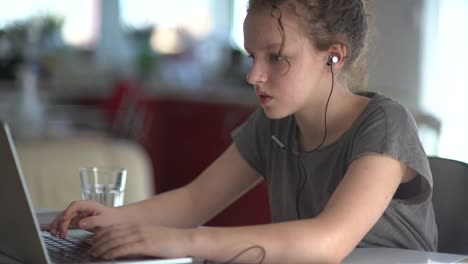 Children,-education-and-distant-learning-concept---teenage-student-girl-in-headphones-with-laptop-computer-at-home.-Cute-girl-listens-attentively,-answers-and-writes-in-a-notebook