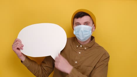 Retrato-del-joven-asombrado-con-máscara-facial-protectora-médica-sosteniendo-y-apuntando-a-la-burbuja-del-habla-en-blanco,-lleva-camisa-y-sombrero-de-gorro,-aislado-en-el-fondo-amarillo-del-estudio.-Concepto-de-estilo-de-vida-de-las-personas