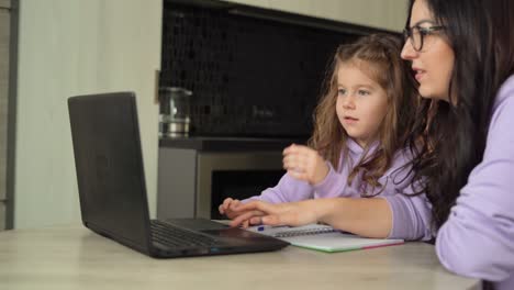 Mamá-e-hija-están-sentadas-en-una-mesa-en-la-cocina.-Mamá-enseña-a-su-hija-pequeña-a-usar-una-computadora-portátil-e-Internet.