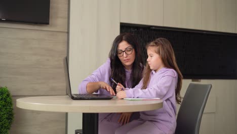 Mamá-feliz-y-la-hija-pequeña-de-preescolar-están-sentados-en-la-mesa-de-la-cocina,-usando-computadora-portátil,-haciendo-la-tarea-a-la-escuela.-concepto-de-educación-a-distancia-en-línea