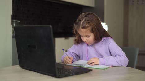 Fernunterrichtskonzept.-eine-kleine-Schülerin-studiert-online-mit-einem-Laptop,-Notizen-in-einem-Notizbuch-machen.-das-Kind-sitzt-am-Tisch