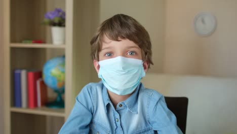 Retrato-del-colegial-niño-niño-con-máscara-médica-protectora-mirando-a-la-cámara,-estudiando-en-casa