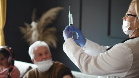 Female-doctor-prepares-a-needle-for-vaccinating-an-older-grandfather-at-home,-vaccinating-the-family-in-the-living-room.