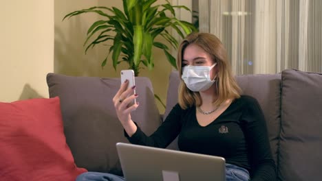 Junge-Türkin-mit-Gesichtsmaske,-die-einen-Videoanruf-auf-ihrem-Smartphone-hat,-während-sie-auf-dem-Sofa-sitzt.-Auf-ihrem-Schoß-befindet-sich-ein-Laptop,-eine-Kaffeetasse-und-ein-Handdesinfektionsbottole-auf-dem-Tisch.