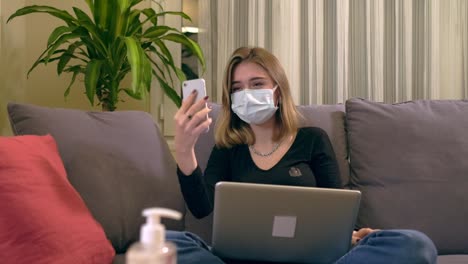 Young-Turkish-woman-with-a-facial-mask,-having-a-video-call-on-her-smartphone-while-sitting-on-the-sofa.-There-is-a-laptop-computer-on-her-lap,-coffee-cup-and-a-hand-sanitizer-bottole-on-the-table.