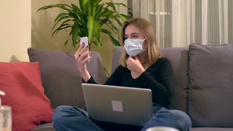 Junge-Türkin-mit-Gesichtsmaske,-die-einen-Videoanruf-auf-ihrem-Smartphone-hat,-während-sie-auf-dem-Sofa-sitzt.-Auf-ihrem-Schoß-befindet-sich-ein-Laptop,-eine-Kaffeetasse-und-ein-Handdesinfektionsbottole-auf-dem-Tisch.