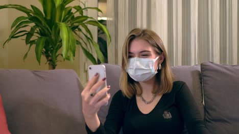 Young-Turkish-woman-with-a-facial-mask,-having-a-video-call-on-her-smartphone-while-sitting-on-the-sofa.-There-is-a-laptop-computer-on-her-lap,-coffee-cup-and-a-hand-sanitizer-bottole-on-the-table.