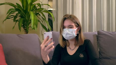 Junge-Türkin-mit-Gesichtsmaske,-die-einen-Videoanruf-auf-ihrem-Smartphone-hat,-während-sie-auf-dem-Sofa-sitzt.-Auf-ihrem-Schoß-befindet-sich-ein-Laptop,-eine-Kaffeetasse-und-ein-Handdesinfektionsbottole-auf-dem-Tisch.