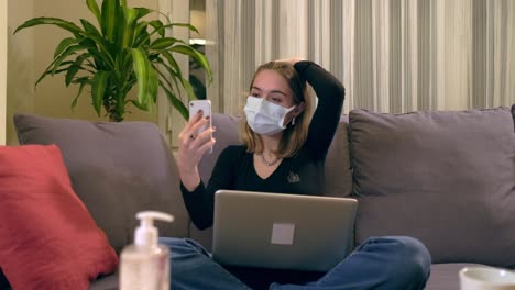 Junge-Türkin-mit-Gesichtsmaske,-die-einen-Videoanruf-auf-ihrem-Smartphone-hat,-während-sie-auf-dem-Sofa-sitzt.-Auf-ihrem-Schoß-befindet-sich-ein-Laptop,-eine-Kaffeetasse-und-ein-Handdesinfektionsbottole-auf-dem-Tisch.