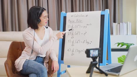 Asiatische-Schule-junge-Lehrerin-arbeiten-von-zu-Hause-lehren-Online-Mathematik-Fach-zu-Studenten-von-zu-Hause-studieren.-Mädchen-zeigt-auf-Whiteboard,-sprechen-Sie-auf-Kopfhörer.-Fernunterrichtsunterricht-während-der-covid19-Pandemie