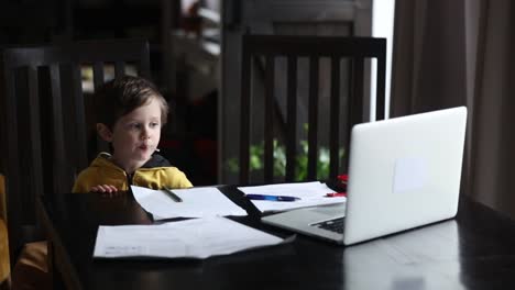 Kleiner-Junge-sitzt-am-Tisch-mit-Laptop-und-Dokumente-für-den-Unterricht-zu-Hause