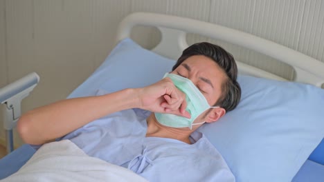 Unwell-young-Asian-male-patient-coughing-while-rest-lying-in-hospital-bed-recovery-after-fell-ill-with-cold-and-flu.-Wearing-face-mask-in-quarantine-for-protection-during-covid-19-wait-for-treatment.