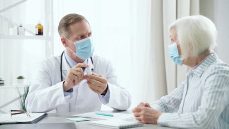 Prevent-getting-sick.-Middle-aged-male-doctor-wearing-mask-consulting-elderly-patient-woman,-holding-and-showing-a-glass-bottle-of-vaccine,-sitting-at-the-table-in-his-office