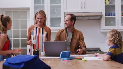 Caucasian-parents-embracing-in-kitchen,-father-working-on-laptop-and-children-doing-school-work