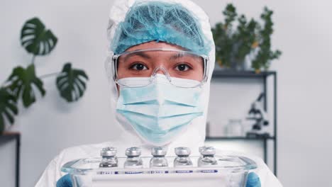 Hope-fighting-coronavirus.-Serious-multiethnic-female-lab-scientist-in-protection-suit-showing-tray-with-vaccine-flasks.