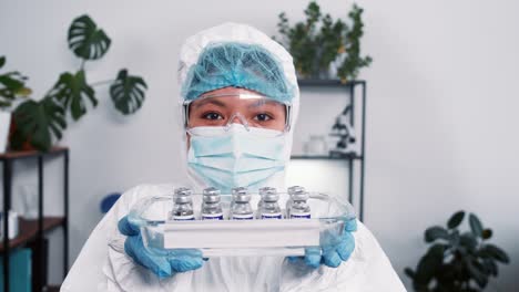 Victory-over-coronavirus.-Cheerful-female-science-lab-worker-in-protection-suit-shows-medical-tray-with-vaccine-flasks.