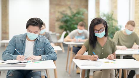 Enthusiastic-learner.-Diverse-students-sitting-at-table-in-university,-wearing-protective-face-mask,-listening-and-making-notes.-Guy-raised-his-hand-during-lecture-in-a-classroom