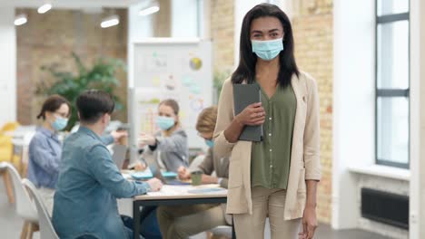 Attraktive-gemischte-Rasse-junge-Frau-trägt-schützende-Gesichtsmaske-Blick-auf-die-Kamera,-stehend-mit-Tablet-PC-in-einem-Klassenzimmer.-Gruppe-unterschiedlicher-Studenten,-die-im-Hintergrund-arbeiten