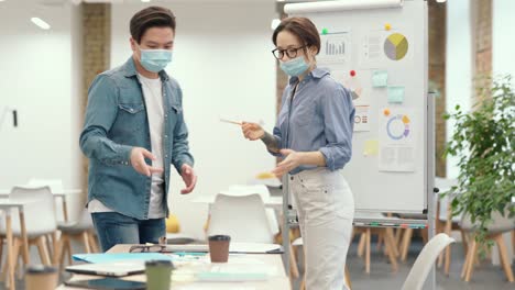 Pandemic-times.-Young-man-and-woman,-students-or-colleagues-wearing-protective-face-masks-greeting-each-other-by-bumping-elbows-and-showing-presentation-during-a-class