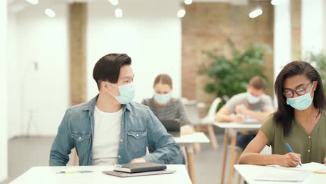 Studying-time.-Male-student-coming-to-a-classroom-with-laptop-and-notebooks,-bumping-elbow-with-his-groupmate-as-a-sign-of-greeting.-Students-wearing-protective-face-mask-writing-test