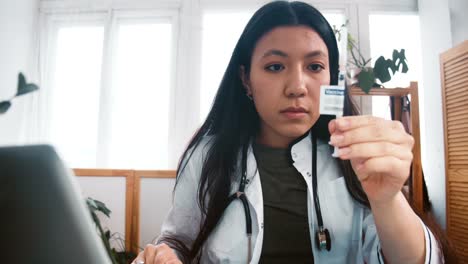 Zoom-in-on-professional-multiethnic-doctor-woman-in-lab-coat-looking-at-new-vaccine-syringe-cure,-checking-with-laptop.