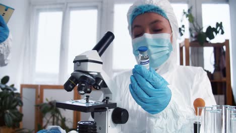 Happy-female-clinic-lab-scientist-in-protection-suit-and-shield-holding-flask-with-new-vaccine.-Hope-in-fighting-disease