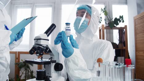 Vaccine-trials.-Professional-female-pharmacy-doctor,-scientist-in-full-protection-suit-holds-flask-with-cure-at-lab.