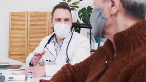 Friendly-young-male-Caucasian-healthcare-professional-in-lab-coat-smiles,-consults-senior-patient-woman-at-clinic-office