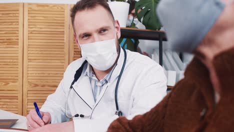 Young-male-Caucasian-healthcare-professional-in-lab-coat-consults-senior-patient-woman-at-clinic-office,-nurse-walks-by.