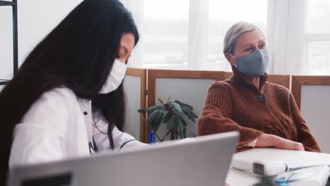 Concepto-de-examen-médico.-Joven-amigable-médico-de-raza-mixta-en-bata-de-laboratorio-consultando-a-la-mujer-cliente-de-pelo-gris-senior.