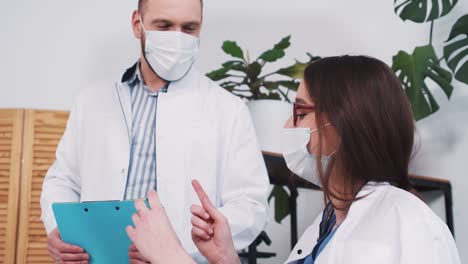 Young-smart-Caucasian-female-pharmacy-doctor-shows-flask-with-vaccine-to-male-colleague-at-light-modern-clinic-office.