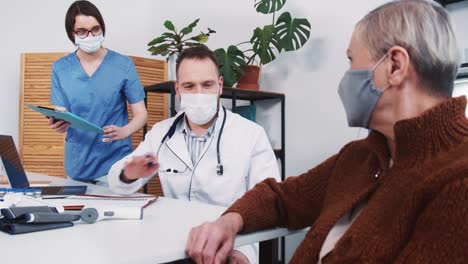 Young-friendly-male-doctor-offers-vaccine-treatment-to-senior-patient-woman-at-clinic-office,-nurse-gives-him-documents.