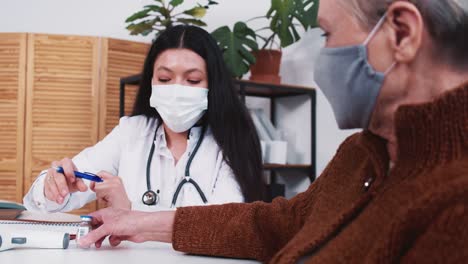 La-doctora-en-farmacia-de-raza-mixta-le-da-frasco-con-medicina-vacunal-a-la-mujer-paciente-caucásica-de-la-tercera-edad-en-el-examen-de-la-clínica.