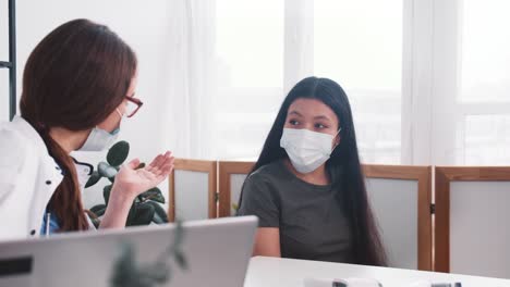 Examen-médico-en-la-clínica.-Joven-médica-amigable-mujer-en-bata-de-laboratorio-consultando-a-la-mujer-paciente-de-raza-mixta-en-máscara-facial.