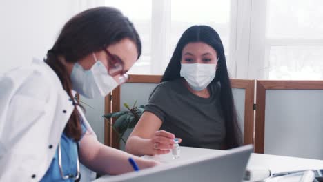 La-joven-doctora-caucásica-consulta-a-una-mujer-paciente-de-raza-mixta-con-máscara,-sosteniendo-el-frasco-de-medicamentos-en-el-examen-médico.
