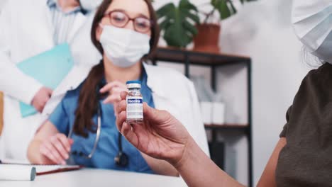 Vaccination-concept.-Young-Caucasian-virologist,-pharmacy-doctor-woman-gives-flask-with-medicine-to-mixed-race-patient.