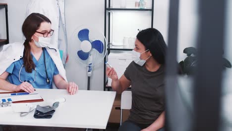 La-joven-doctora-en-farmacia-caucásica-da-frasco-con-medicina-vacunal-a-una-mujer-paciente-de-raza-mixta-en-un-examen-médico.