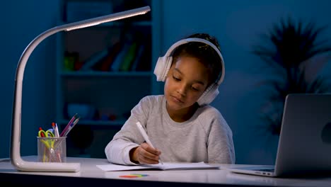 A-girl-in-headphones-takes-notes-while-listening-to-an-audio-lesson,-looks-at-the-laptop,-nods-her-head.-A-girl-sits-at-a-table-under-the-light-of-a-table-lamp.-Close-up.-Slow-motion-ready-59.97fps