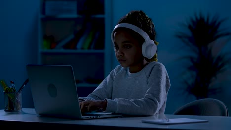 A-serious-African-American-girl-wearing-big-white-headphones-sits-at-a-table-in-a-dark-room,-typing-on-her-laptop.-The-concept-of-distance-learning.-Close-up.-Slow-motion-ready-59.97fps