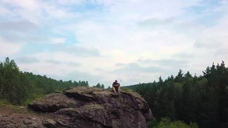 A-sniper-with-a-rifle-sits-on-top-of-the-mountain-and-inspects-the-territory.-View-from-the-drone.-the-camera-moves-from-the-soldier,-revealing-a-view-of-the-landscape