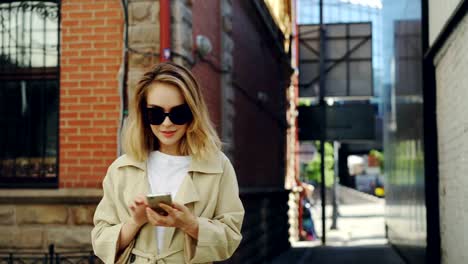 Beautiful-young-lady-is-holding-smartphone-and-using-it-walking-along-street-in-modern-city.-Technology,-beautiful-happy-people-and-youth-lifestyle-concept.