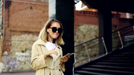 Pretty-girl-is-listening-to-music-through-earphones-using-smartphone-and-dancing-walking-in-the-street-and-enjoying-rhythm-and-modern-city.-Girl-is-wearing-sunglasses-and-coat.