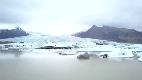 DRONE-aerial-view-of-woman-contemplating-glacier-lagoon-in-Iceland-standing-on-lake-shore