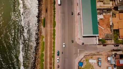 Drone-flying-along-coast-line-street-in-Colombo,-Sri-Lanka.-Aerial-shot-of-cars,-buildings,-ocean-waves-and-railroad