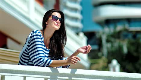 Feliz-sonriente-mujer-turista-vestido-rayas-poner-manos-en-la-barandilla-blanca-y-gafas-de-sol