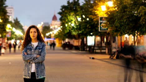 Time-lapse-of-unhappy-girl-standing-alone-on-beautiful-pedestrian-street-late-in-the-evening-and-looking-at-camera-when-people-are-whizzing-around.-Society-and-loneliness-concept.